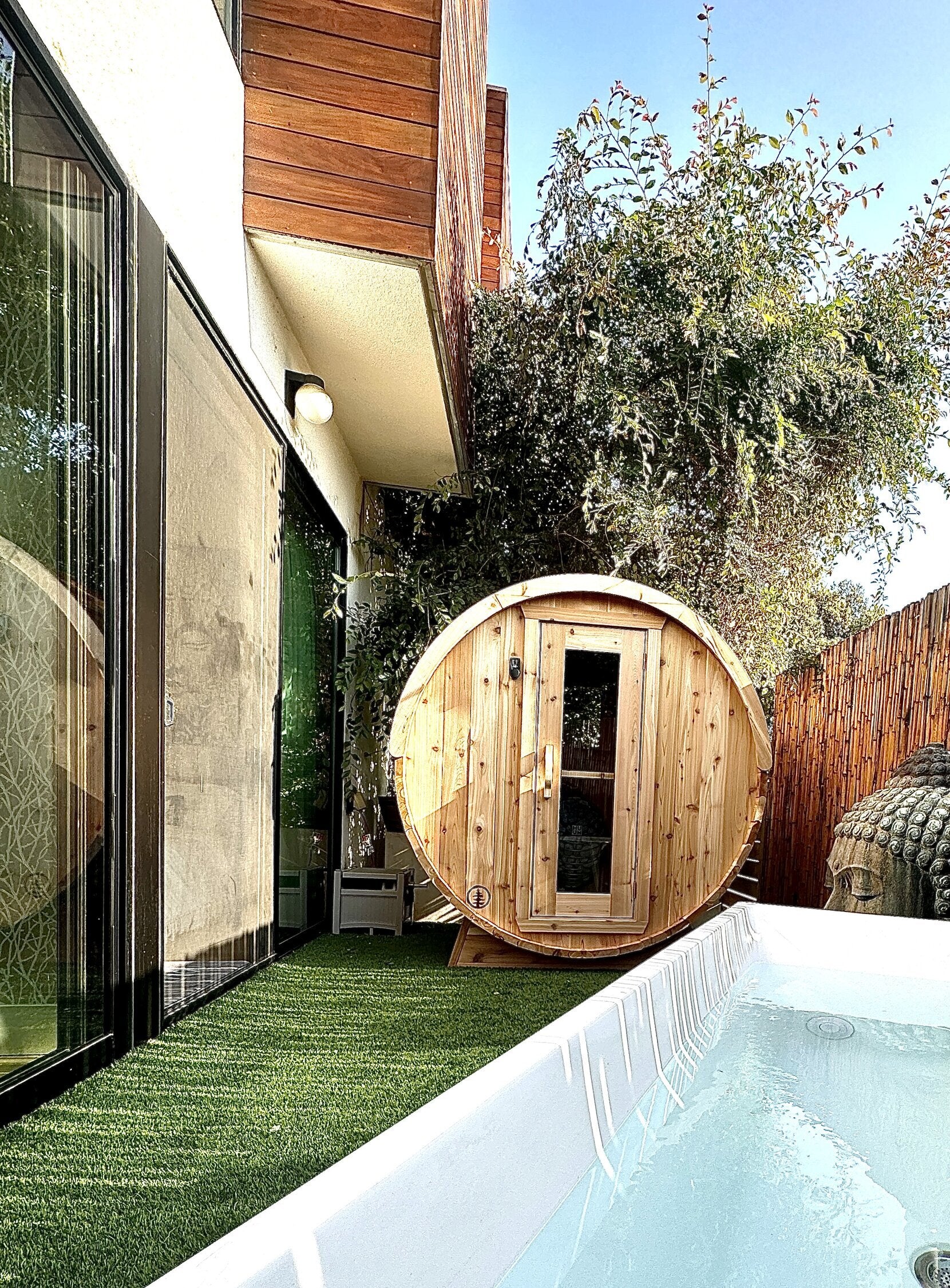 Cedar barrel sauna on artificial turf between a modern house and a bamboo fence, with a white tub in front