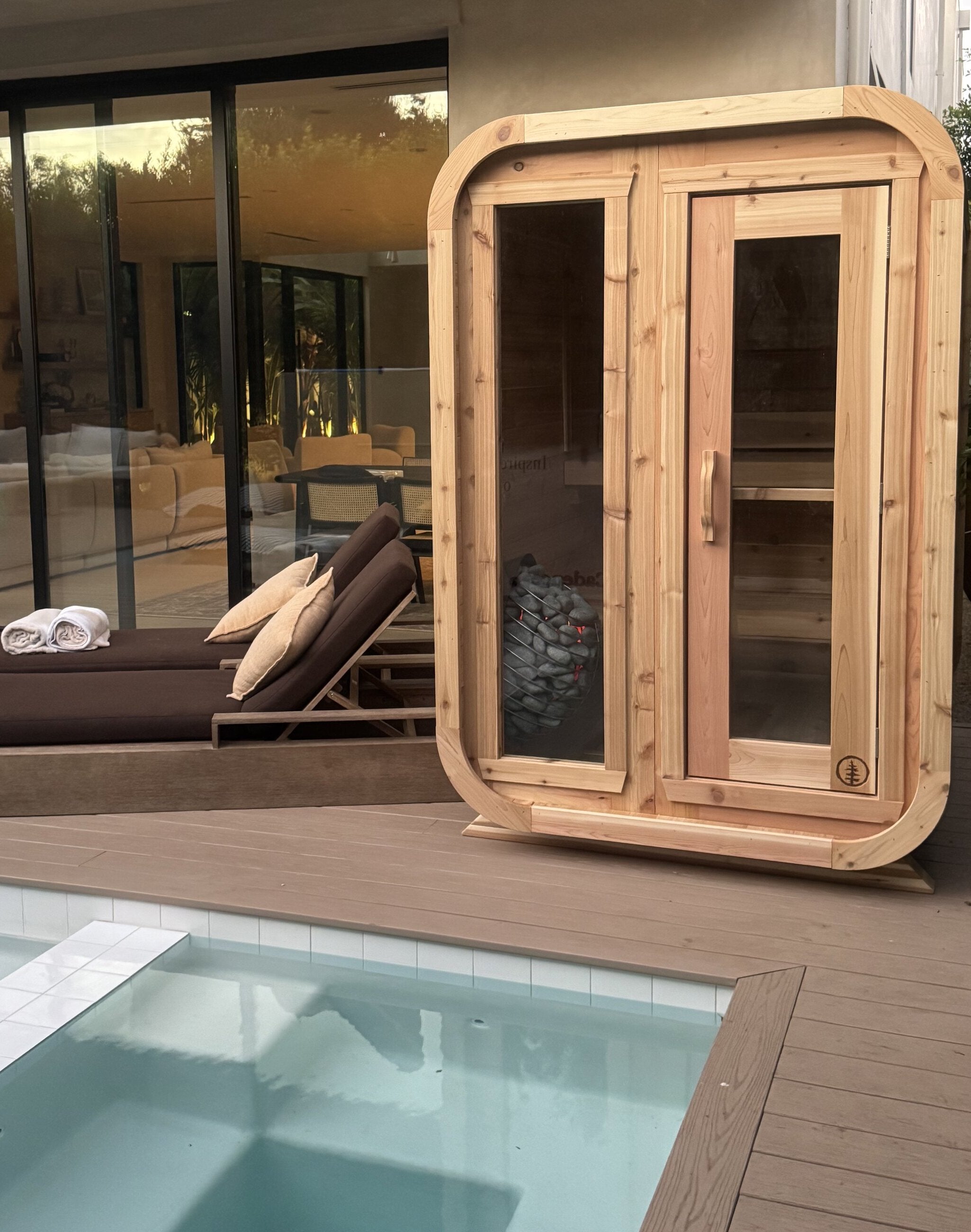 Cedar cabin sauna with glass panels and a glowing heater, beside a small pool and lounge chairs at a modern home
