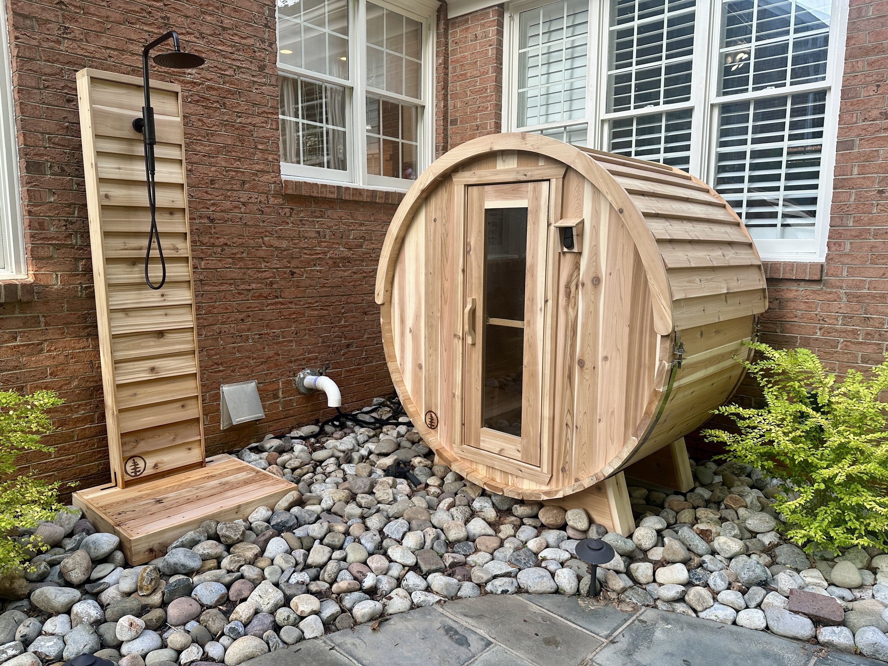 Cedar barrel sauna and cedar outdoor shower on a river-rock bed beside a brick house