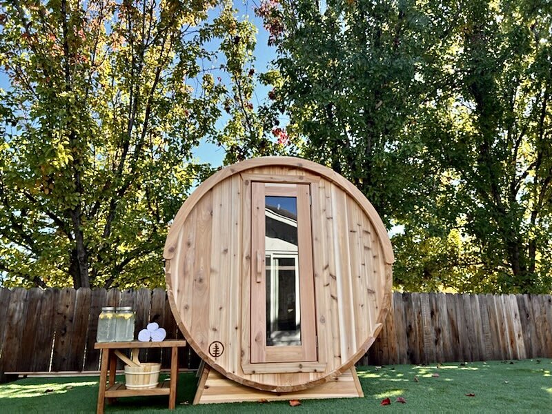 Cedar barrel sauna on artificial turf with a side table of drinks, towels and a cedar bucket under autumn trees