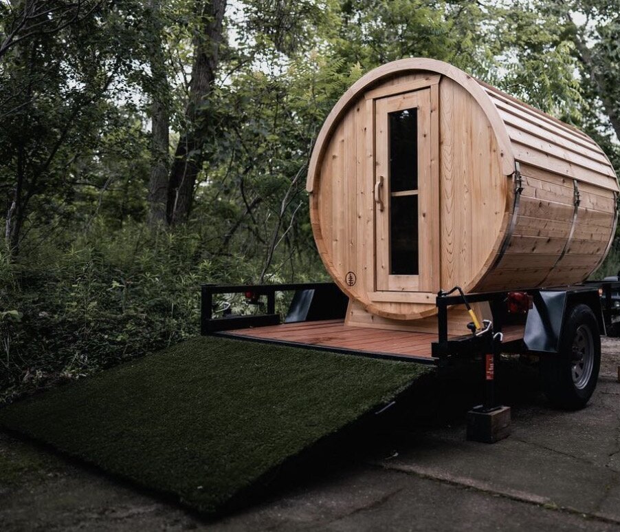 Cedar barrel sauna mounted on a flatbed trailer with a turf-covered ramp, parked at the edge of a forest
