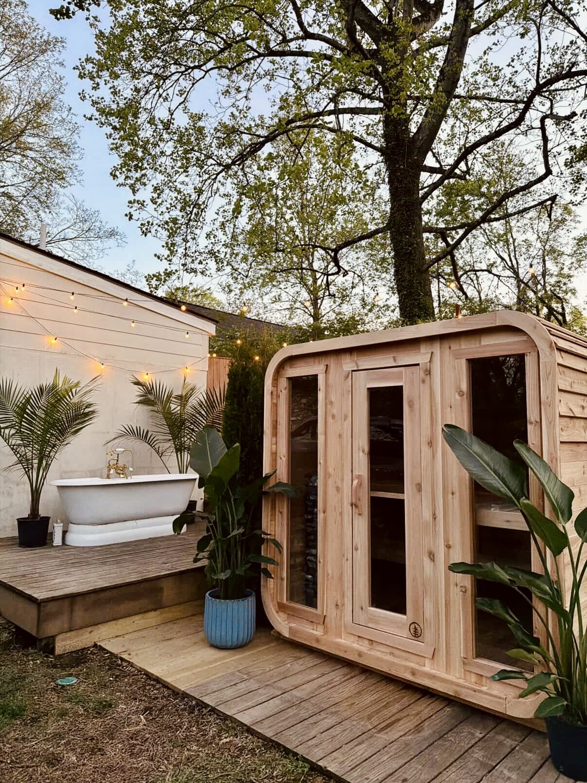 Cedar cabin sauna on a wooden deck at dusk, with string lights, potted palms and a white clawfoot tub