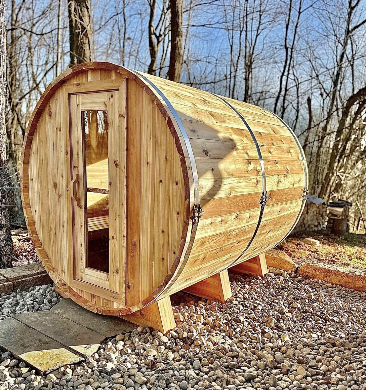 Cedar barrel sauna on a river-rock pad in bare winter woods, lit by warm low sun