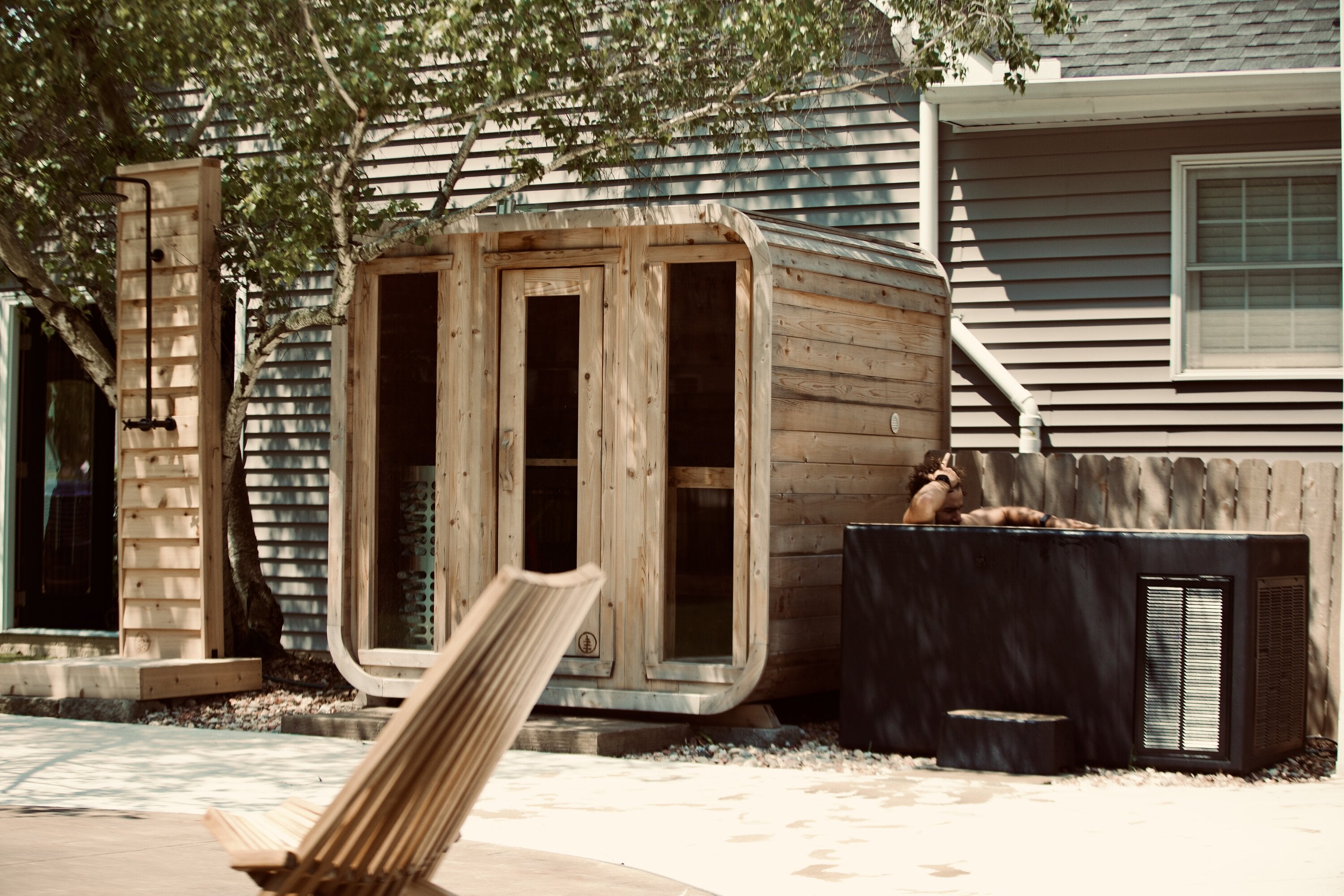 Cedar cabin sauna with three glass front panels, a cedar outdoor shower and a man in a black cold plunge on a patio