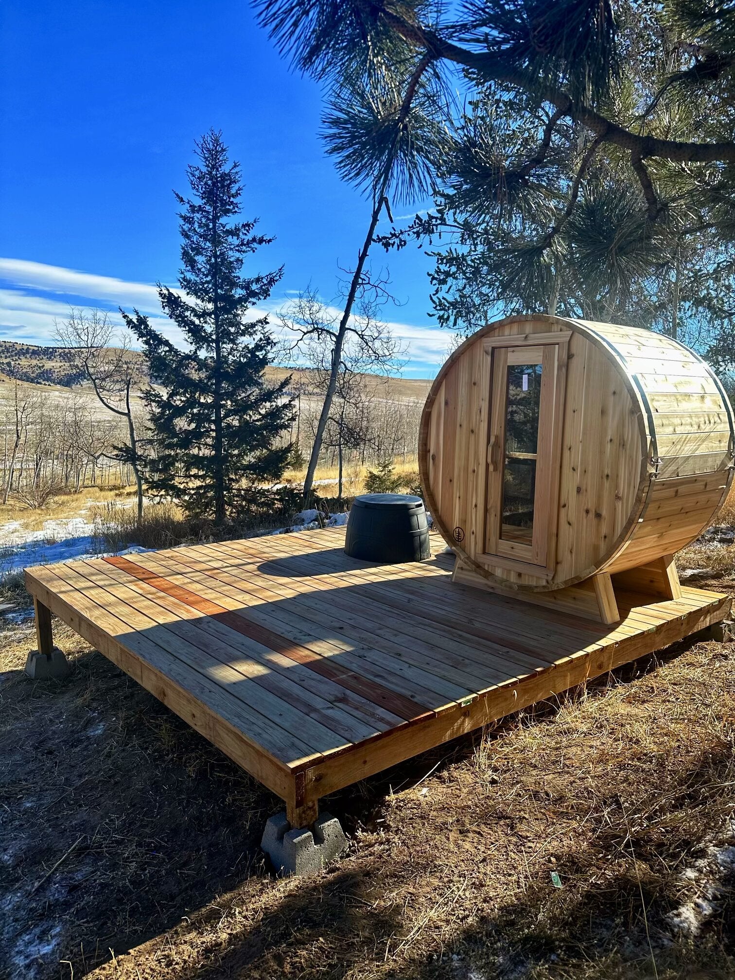 Cedar barrel sauna on a new wood deck among pines, overlooking open hills with patches of snow