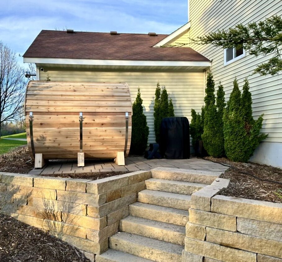 Long cedar barrel sauna seen side-on on a paver pad behind a house, above stone steps and a retaining wall