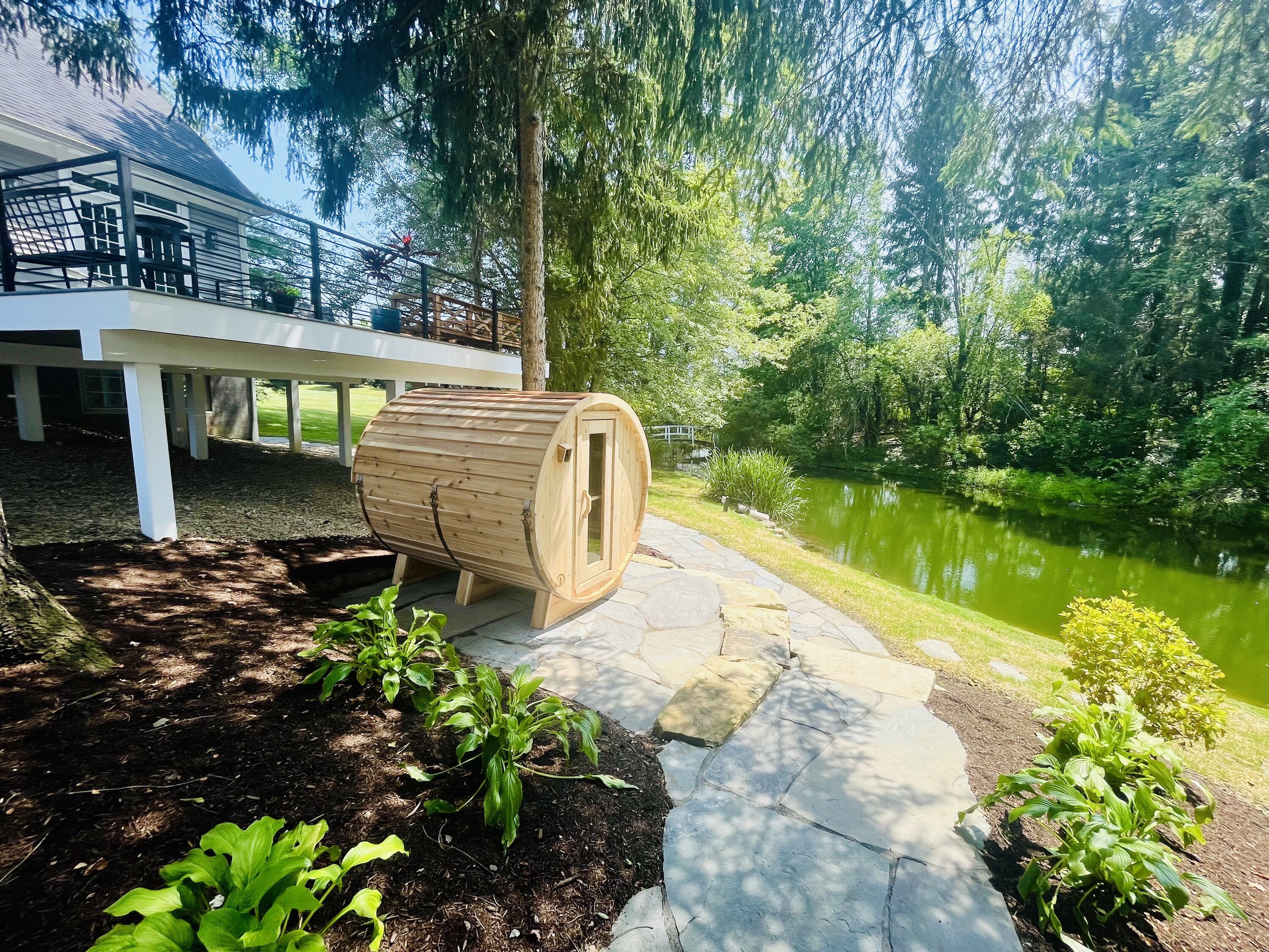 Cedar barrel sauna on a flagstone path beside a calm pond, beneath a raised house deck