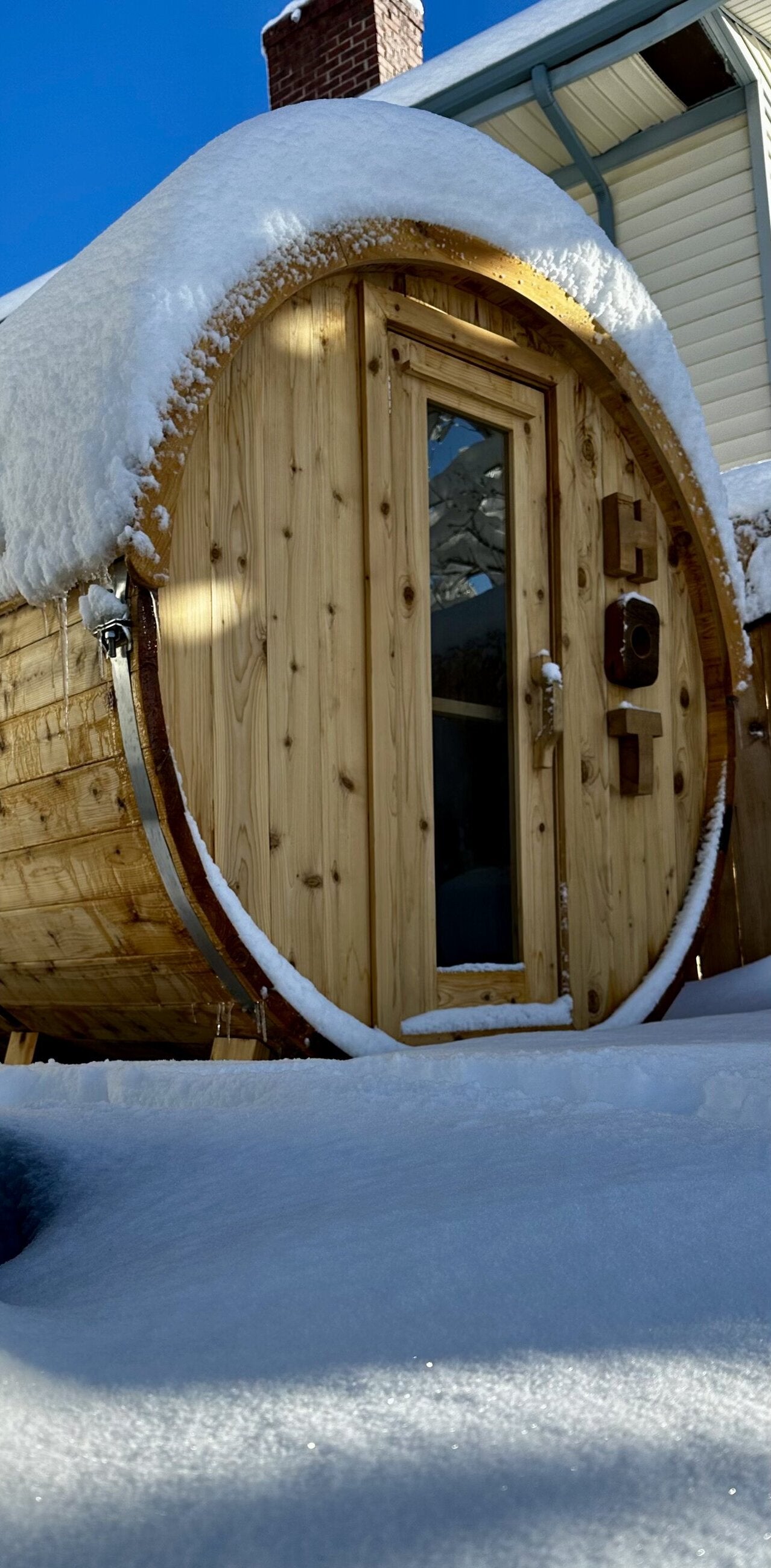 Cedar barrel sauna heaped with fresh snow under a clear blue sky, seen from ground level