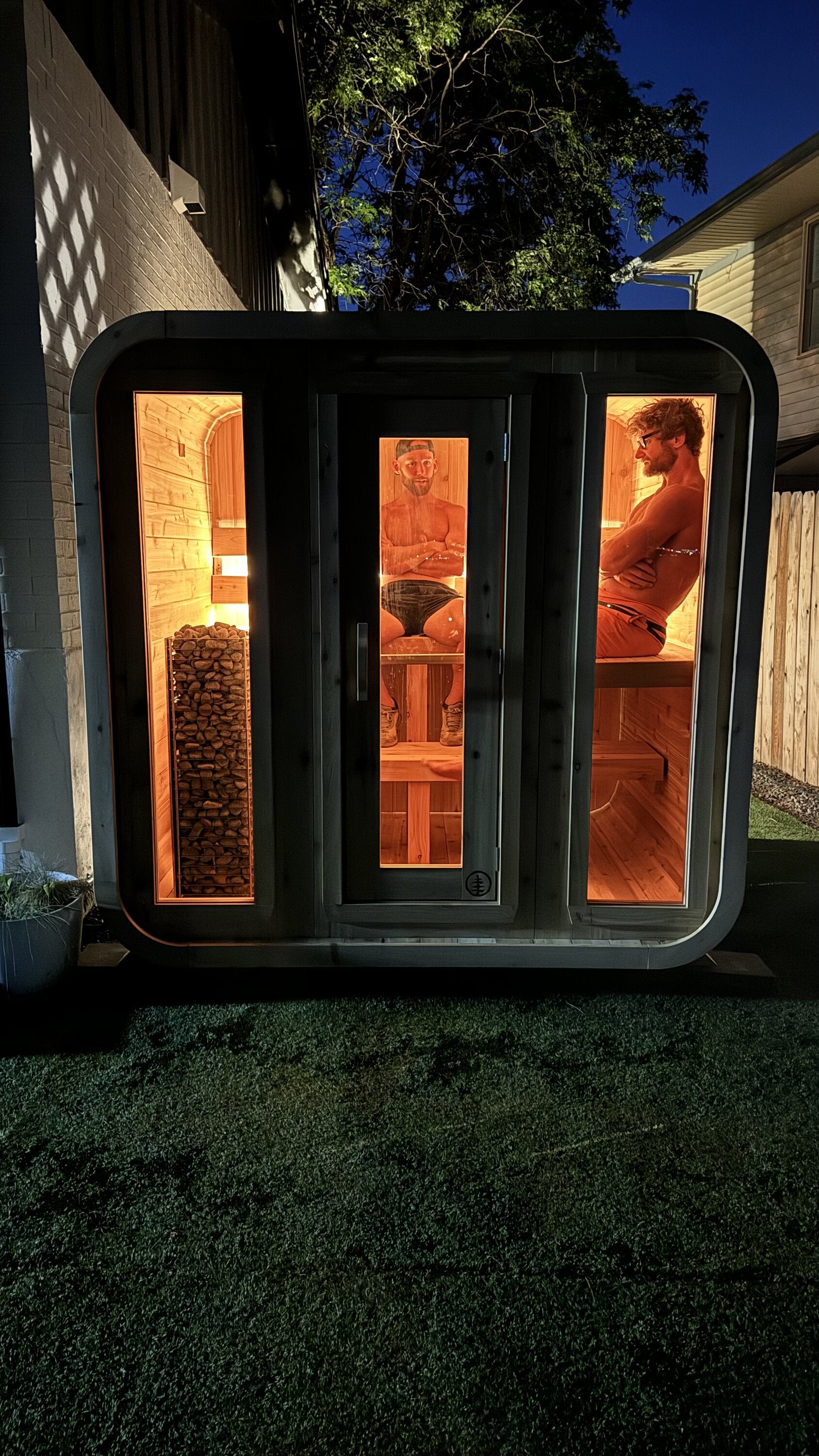 Cabin sauna glowing warm orange at night with two people relaxing inside, seen through three glass panels