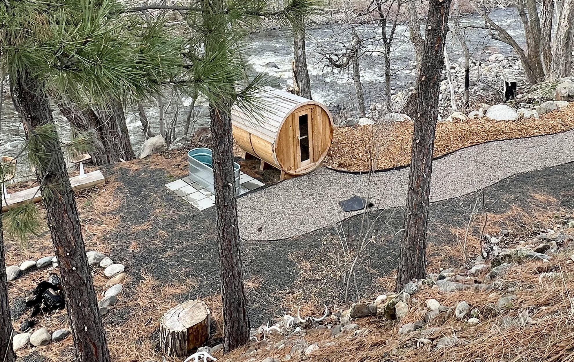 Long cedar barrel sauna and a stock tank plunge beside a gravel path and a rushing river, seen through pines