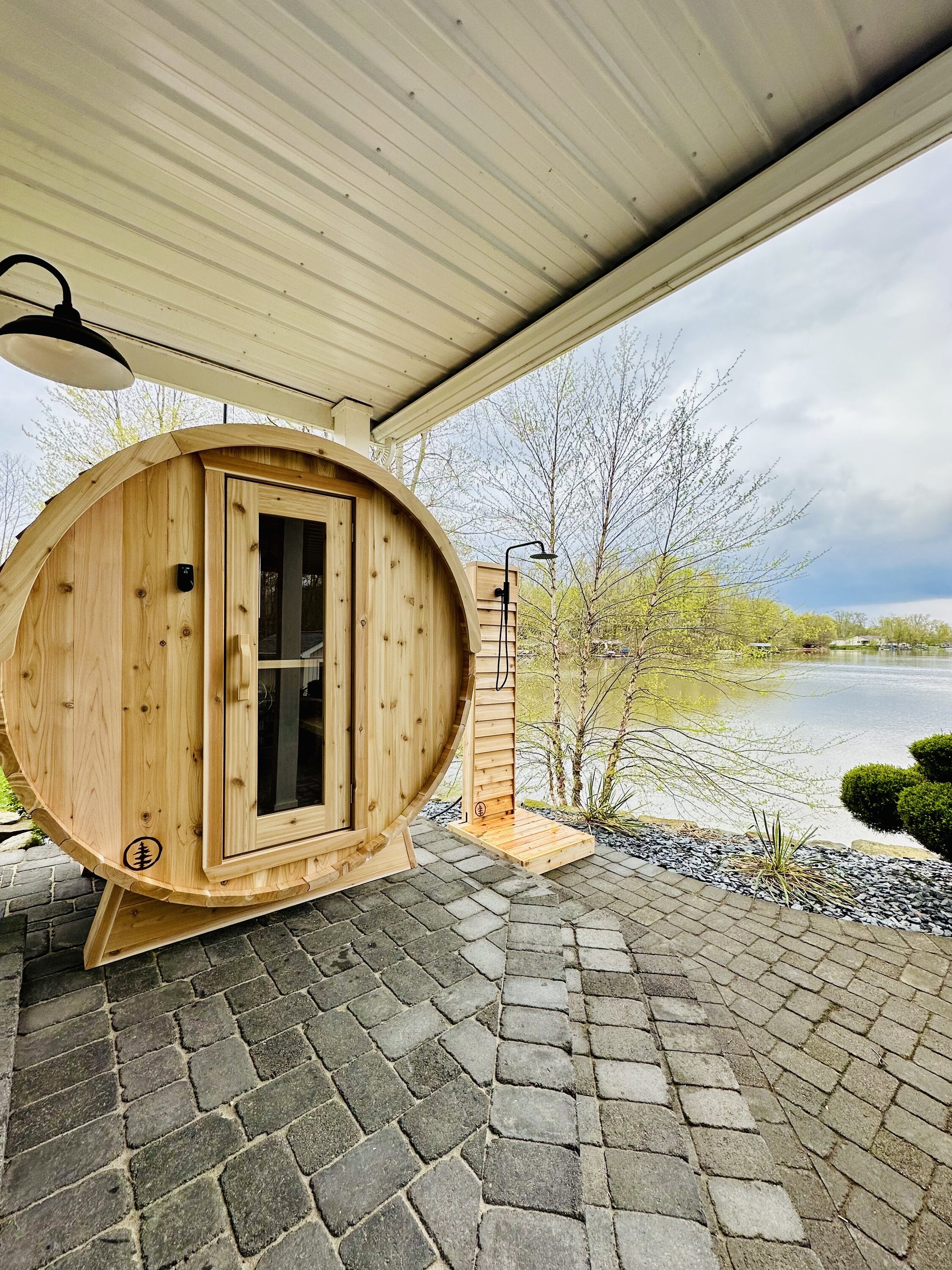 Cedar barrel sauna and cedar outdoor shower on a covered paver patio beside a lake with budding trees