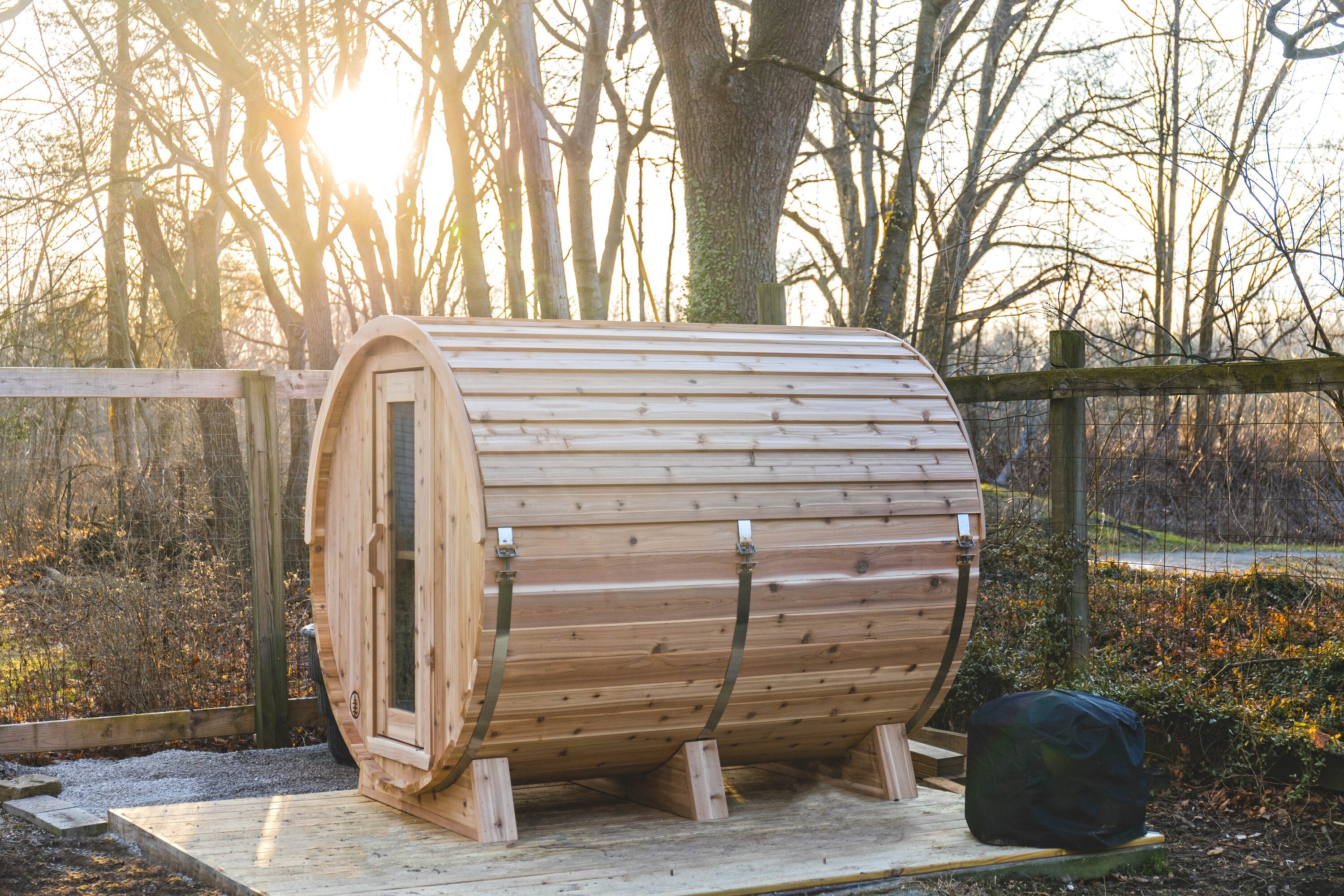 Cedar barrel sauna on a wooden platform at the edge of the woods, sun glowing through bare trees