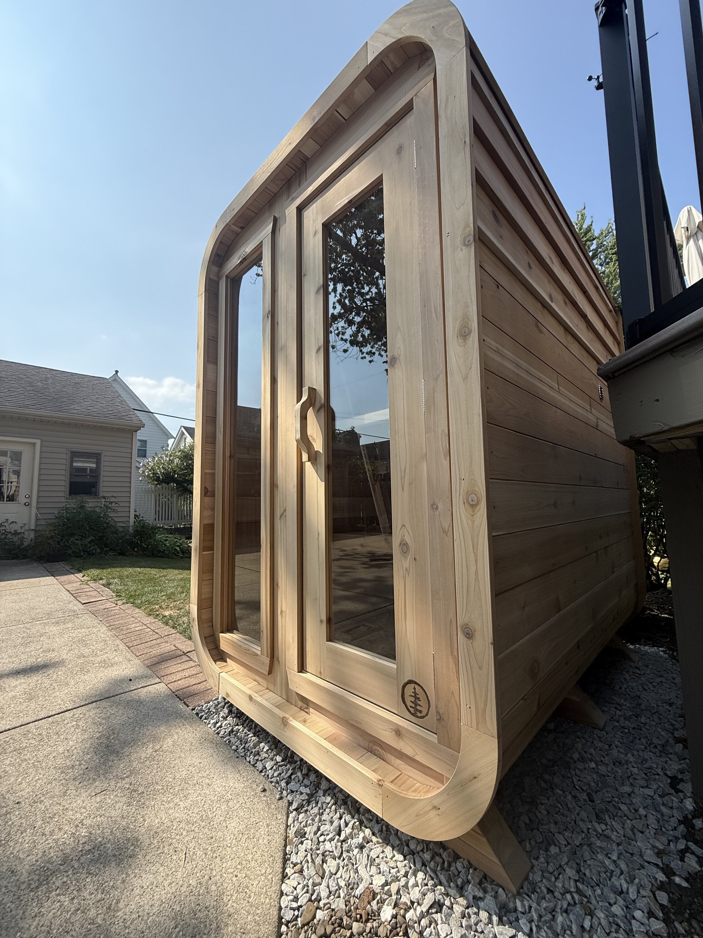 Low-angle view of a rounded cedar cabin sauna with two tall glass panels on a gravel pad under a blue sky
