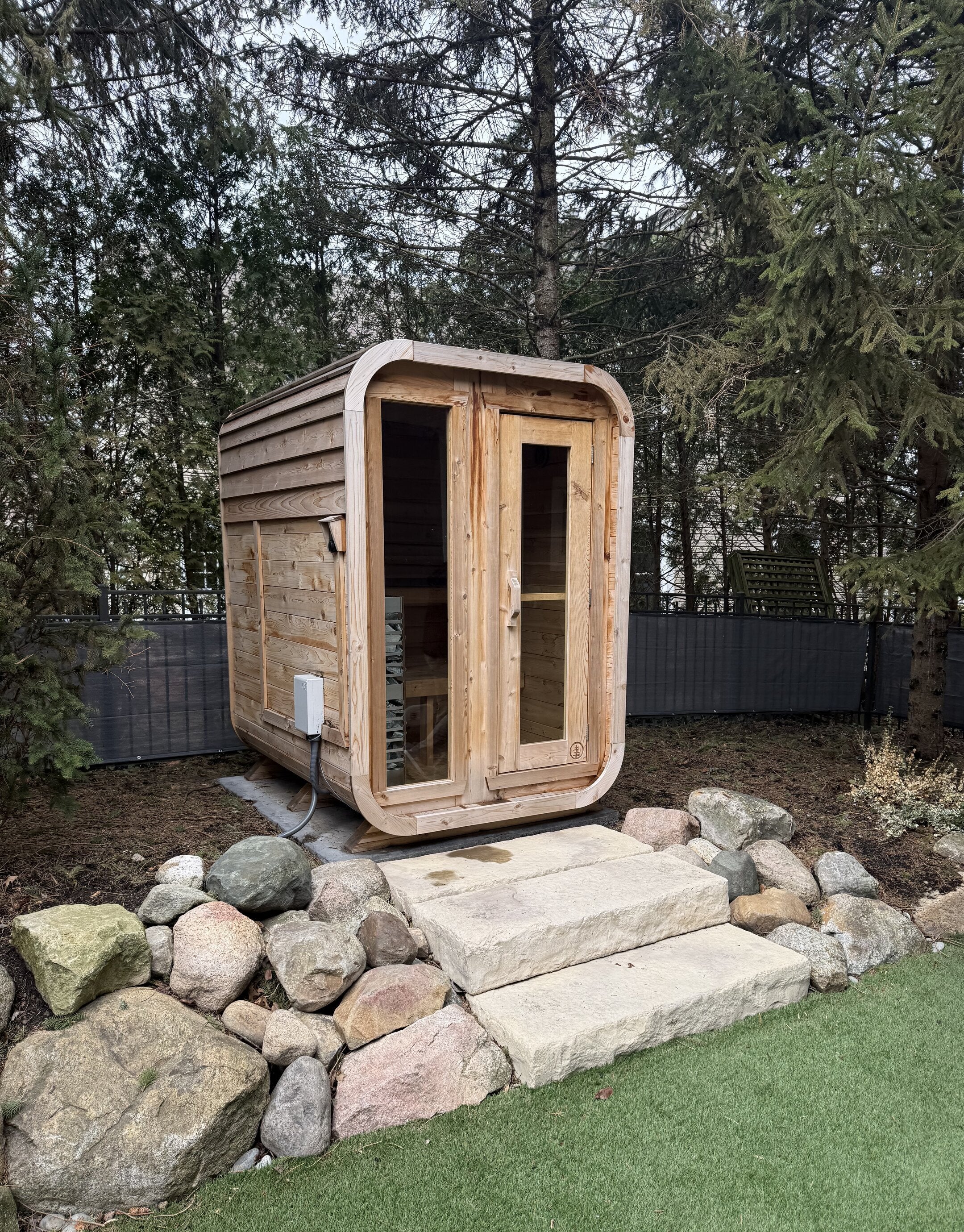 Compact cedar cabin sauna with a glass front among evergreens, reached by stone slab steps from the lawn