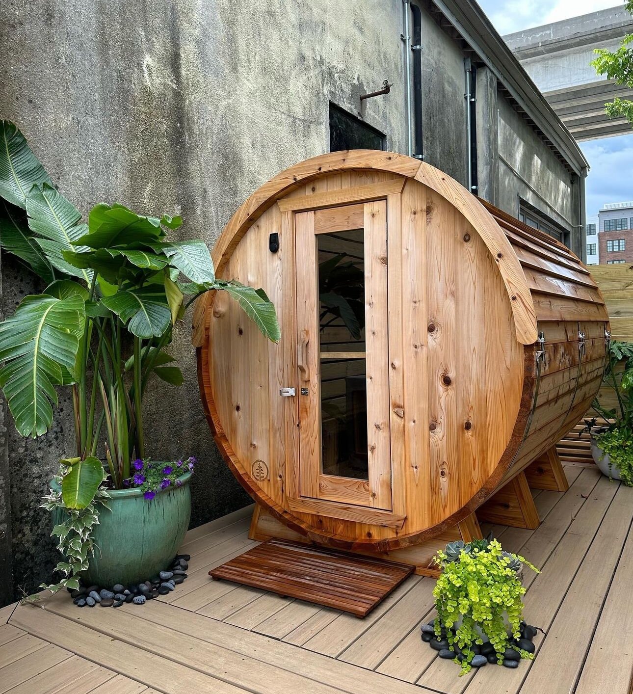 Cedar barrel sauna on a composite deck framed by tropical plants against a concrete wall