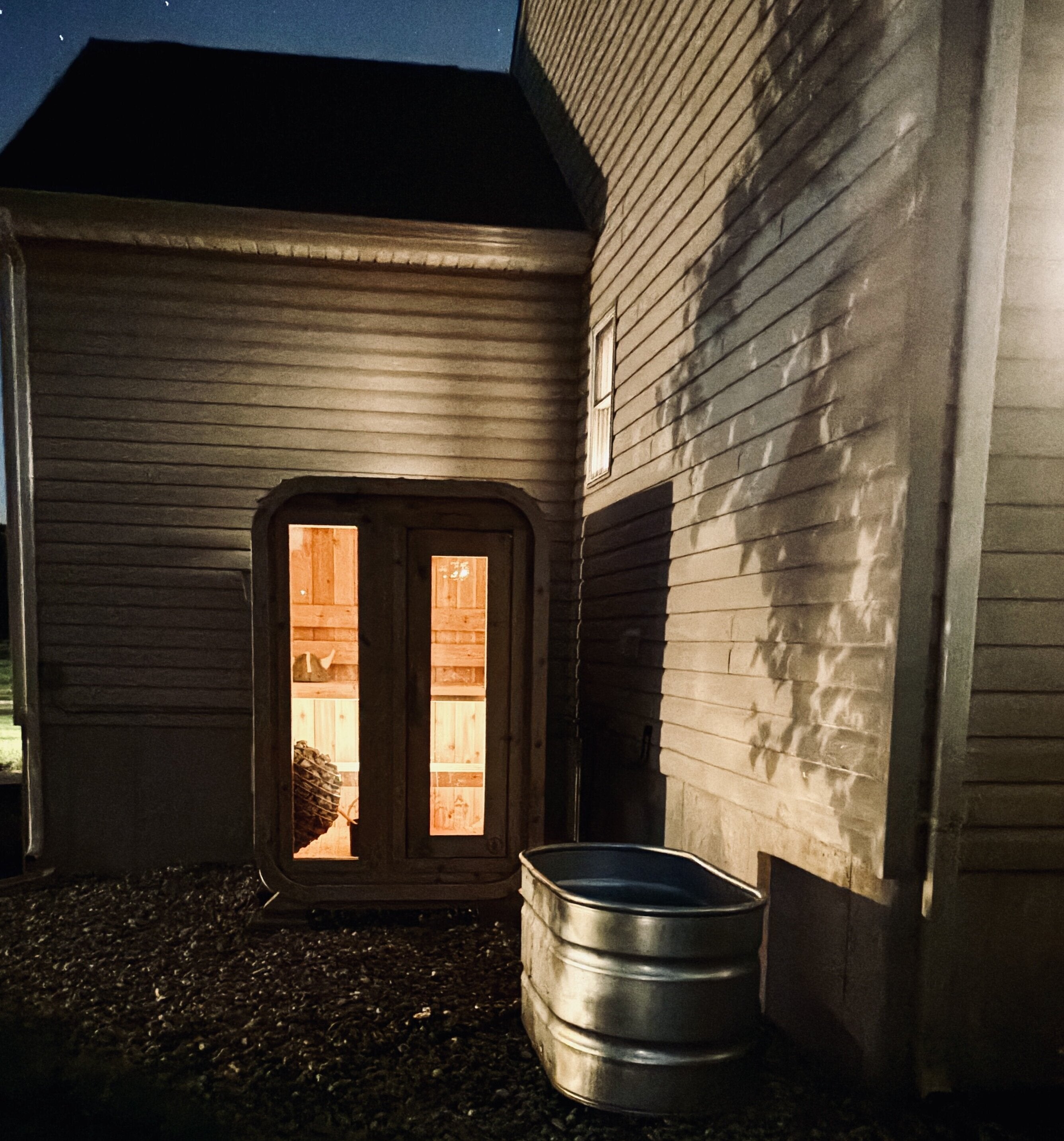 Cabin sauna glowing warm at night against a house wall under the stars, with a metal stock-tank plunge beside it
